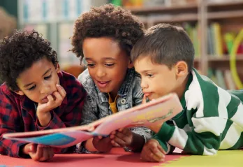 Three kids laying on the floor sharing a book and reading it.