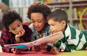 Three kids laying on the floor sharing a book and reading it.